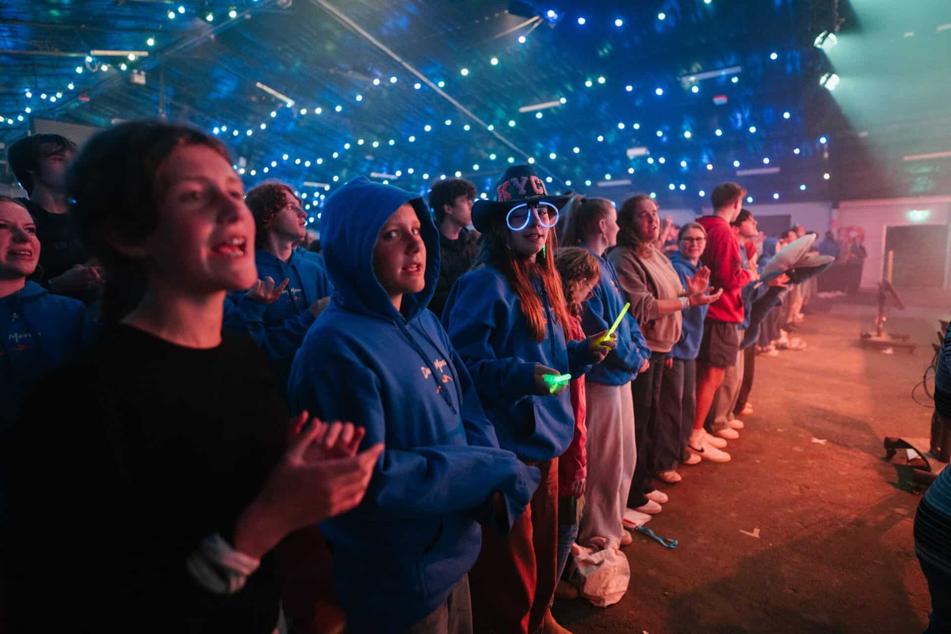 Young people attending a Katoomba Christian Convention event, standing in a line with glow sticks, under vibrant stage lighting, engaging in worship or activities.