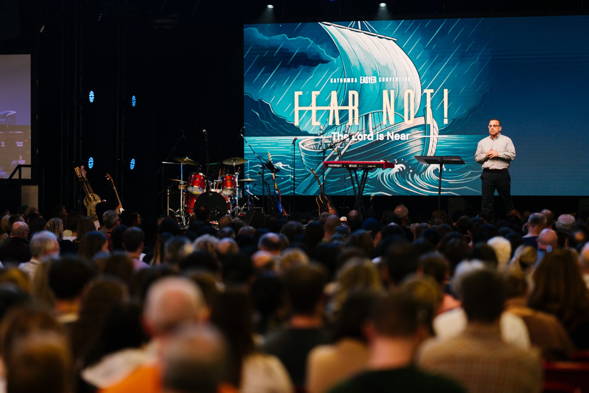Speaker on stage at Katoomba Easter Convention with large screen displaying 'Hear and Now' and a crowd in attendance.