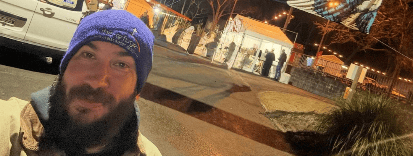 A man with a beard and blue beanie smiling outdoors at night during the Katoomba Christian Convention, with tents and event lighting in the background.