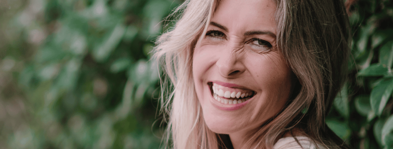 A woman with a bright smile enjoying a moment of happiness in a lush green environment, symbolising faith, hope, and the grace of God in motherhood and life’s journey.