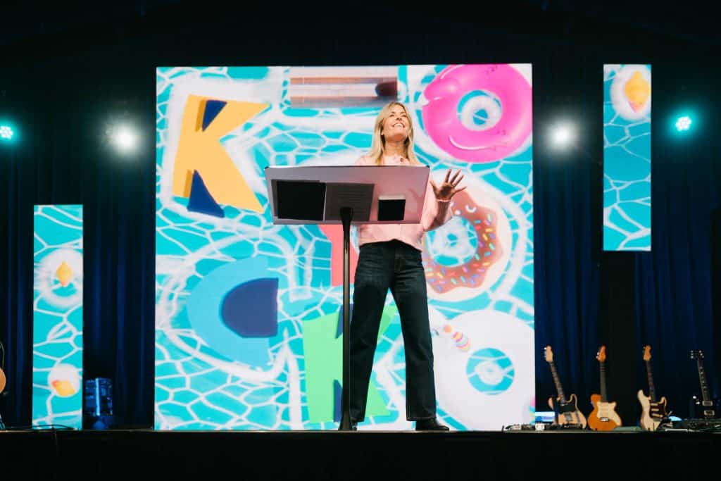 An inspiring speaker at KYCK 26 youth conference, delivering a message of faith and hope on stage with vibrant background visuals.