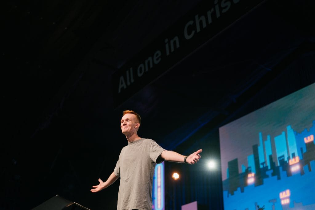 A speaker at KYCK 26 conference on stage with a large screen behind, delivering a message about unity in Christ.