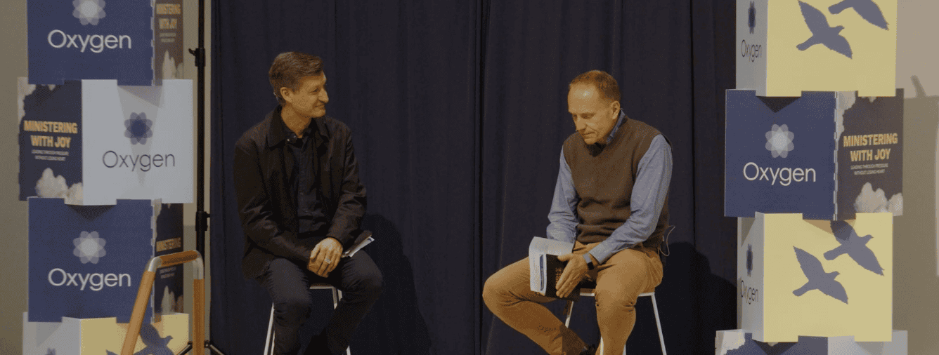 Two men engaged in a discussion on stage at the Oxygen 'Ministering with Joy' Conference, with banners displaying the Oxygen logo and conference theme in the background.