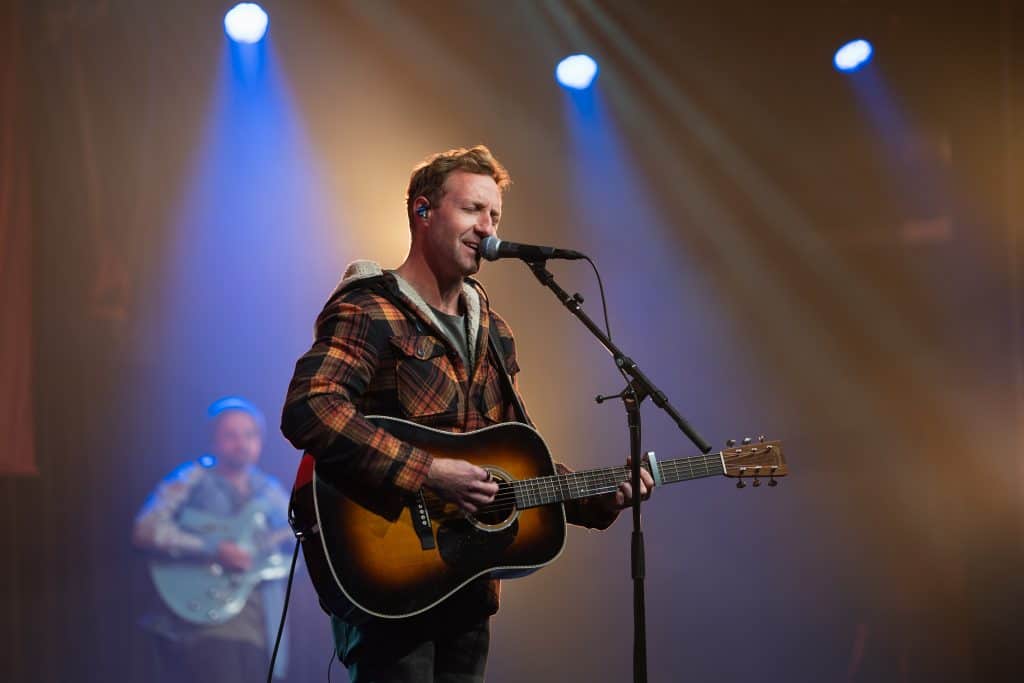 Male musician singing and playing acoustic guitar on stage at BASECAMP 2026 event, with another musician in the background, under stage lighting.