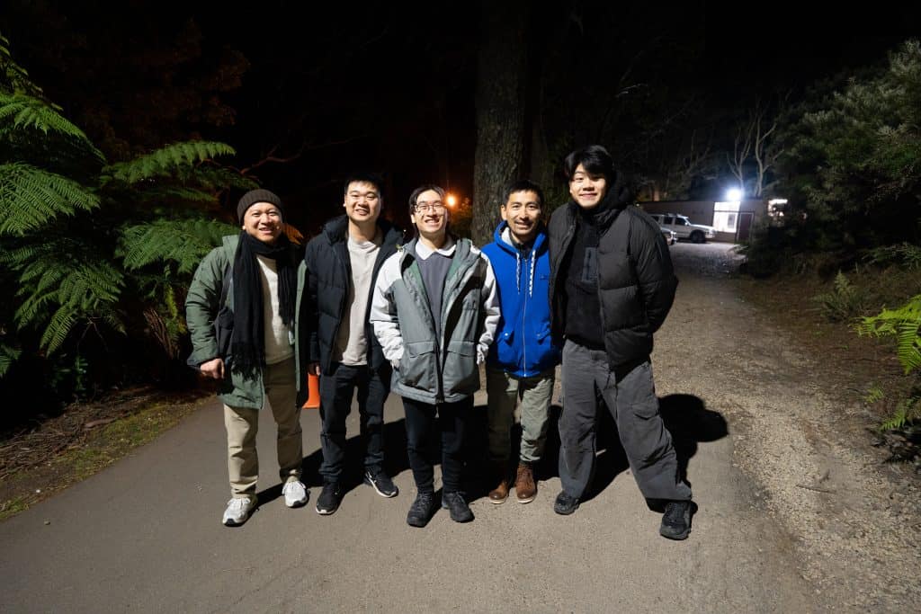 Group of five men standing outdoors at night, smiling, with trees and a vehicle in the background, during BASECAMP 2026.