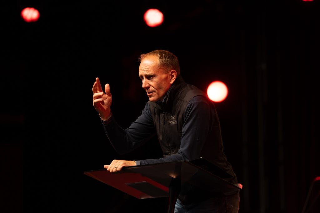 A man speaking passionately at BASECAMP 2026 event, emphasizing resolute faith and leadership among men, with stage lighting highlighting the speaker.