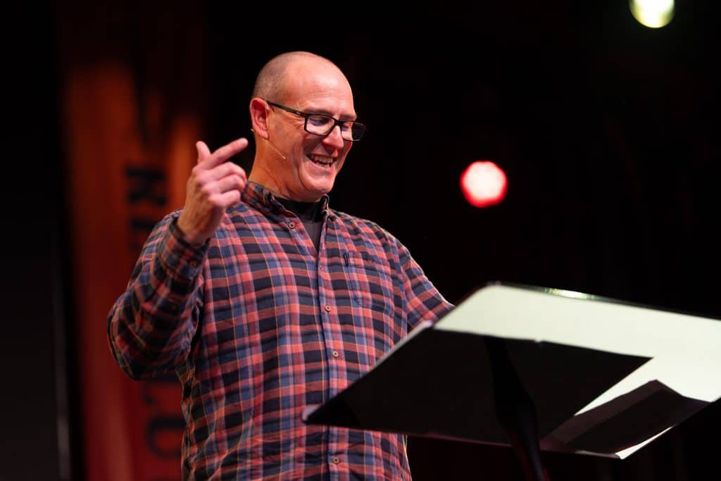 A smiling man in a checkered shirt speaking at a Christian event, with a music stand in front of him and stage lights in the background.