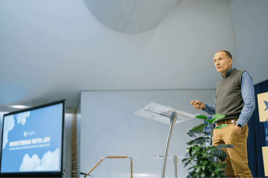 A speaker presenting at the Katoomba Christian Convention during the 'Ministering with Joy' conference, with a presentation slide visible in the background.