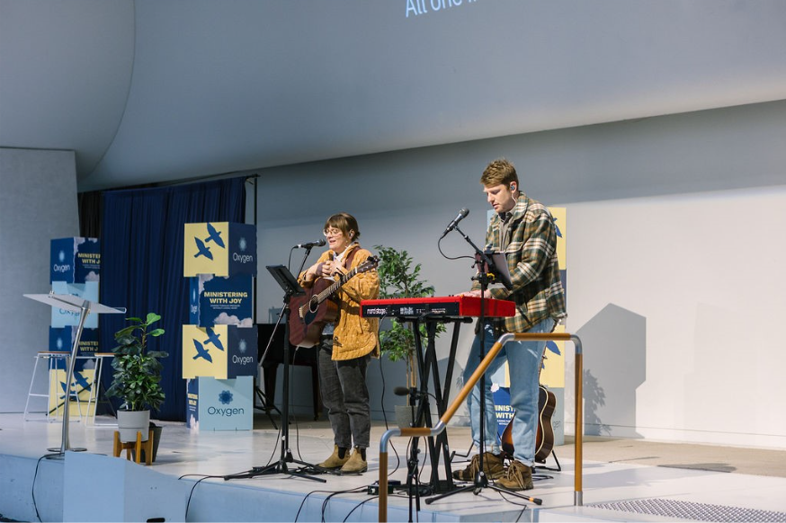 Musicians performing at the Oxygen 'Ministering with Joy' Conference, with a backdrop featuring the event's branding and a stage setup.