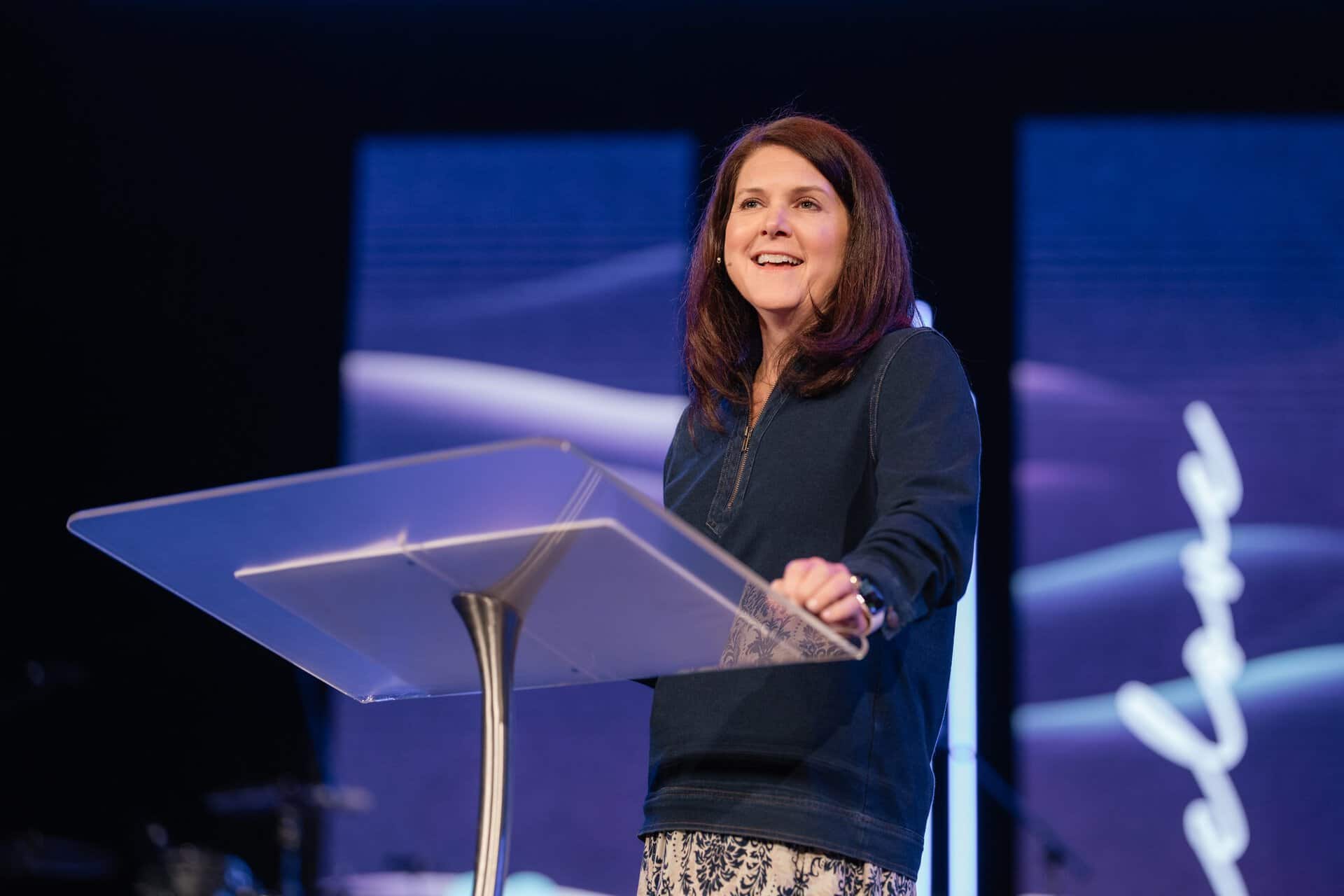 Female speaker at Katoomba Christian Convention delivering a speech on stage with a clear podium and large screens in the background.
