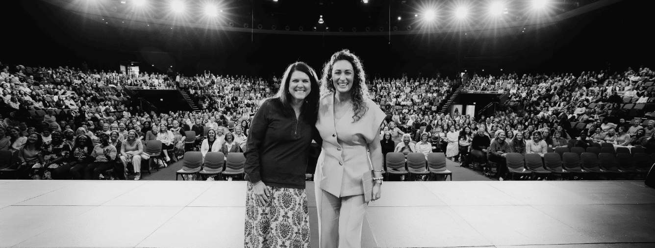 Two women smiling on stage at the OneLove women's conference with a large audience in the background, promoting trust and spiritual growth.